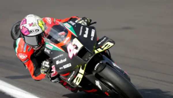 Piloto de MotoGP inclinado en una curva, con su casco blanco y rosa y la moto negra con detalles amarillos.