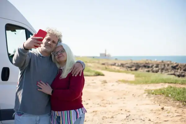 Para singli po 50-tce, którzy szukają przygód, idealne są wycieczki kamperem. Para robi sobie selfie na tle morza.