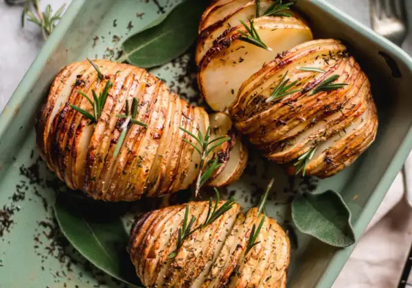 Pommes de terre Hasselback dorées et croustillantes, garnies de romarin frais et d'herbes, dans un plat vert.