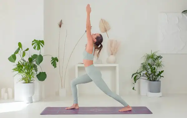 Donna in posa yoga, che mostra i benefici di una pratica regolare. L'ambiente è luminoso e pieno di piante, evocando calma e benessere.