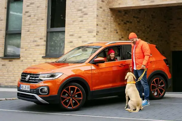 Pomarańczowy mini suv z kobietą w środku i mężczyzną z psem na smyczy.