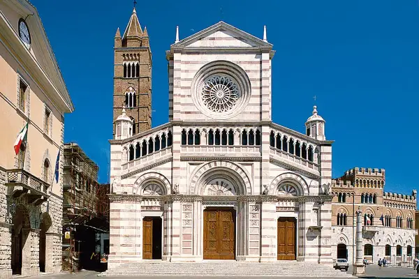 Il Duomo di Grosseto, con la sua facciata a strisce bianche e rosa, domina la piazza. Accanto, un campanile medievale si staglia contro il cielo azzurro.