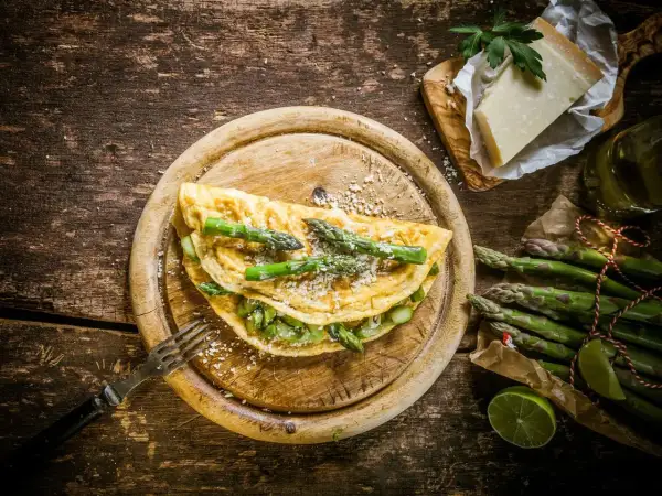 Pyszny omlet ze szparagami, posypany parmezanem, czeka na widelcu. Obok świeże szparagi i kawałek sera.