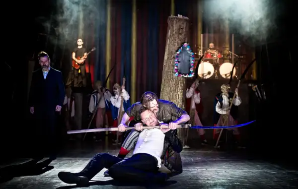 Jan Klata spektakle: dramatyczna scena z kosami i muzyką na żywo. Dwóch mężczyzn walczy na scenie, jeden klęczy, drugi go dusi.