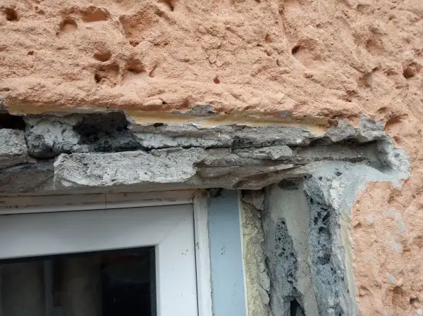 Uszkodzony montaż nadproża w starym domu, widoczne zbrojenie i luźne elementy betonowe nad oknem.