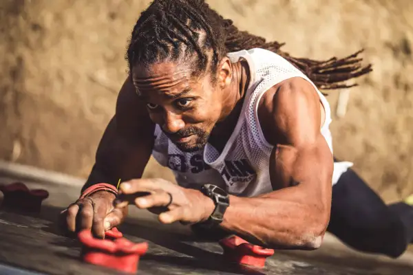 Man with dreadlocks on a climbing wall, his forearm punch showing determination as he grips the holds.