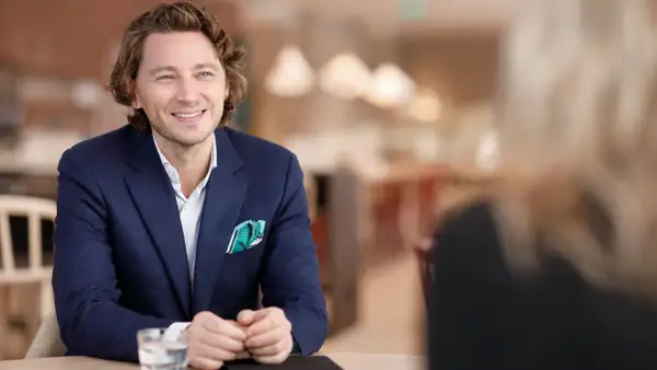 Rasmus Nerman, klädd i en mörkblå kavaj, ler och tittar åt sidan under ett samtal vid ett bord.