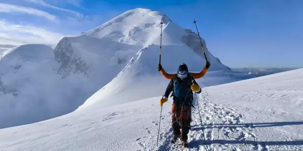 Un alpiniste victorieux lève ses bâtons sur fond de Mont Blanc. L'ascension est réussie, la neige brille sous le ciel bleu.