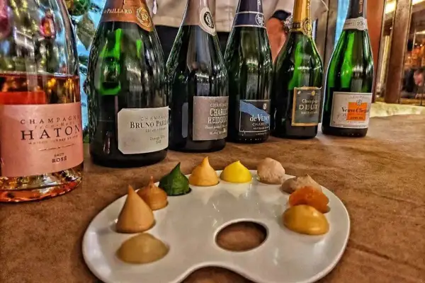 Bottiglie di champagne, tra cui Veuve Clicquot e Bruno Paillard, disposte su un tavolo con una tavolozza da pittore piena di dolcetti colorati, un'ode al gusto e all'arte del cote de bar.