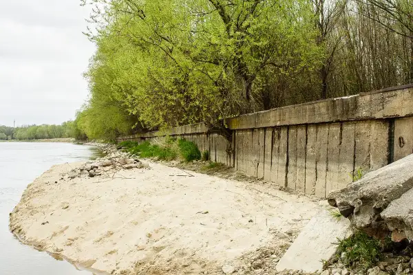 Betonowa opaska na rzece chroni brzeg przed erozją. Wiosenne drzewa tworzą zieloną ścianę nad wodą.