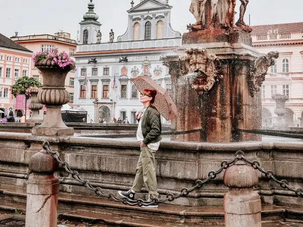 Regenschirm-Schwung am Brunnen in Budweis Stadt. Die Frau genießt die frische Brise, während die historische Kulisse im Hintergrund erstrahlt.