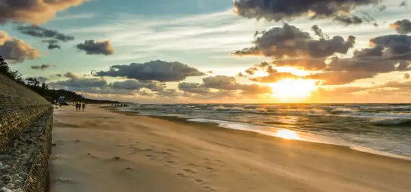Zachód słońca nad Bałtykiem, plaża i falochron. W oddali widać ludzi spacerujących i kitesurferów. To idealne miejsce na podziwianie atrakcji Jastrzębiej Góry i okolic.