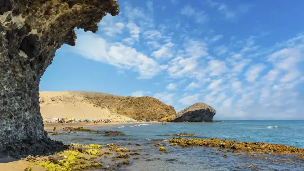 Vista de la playa de San José con rocas cubiertas de algas en primer plano y gente disfrutando del sol bajo sombrillas.