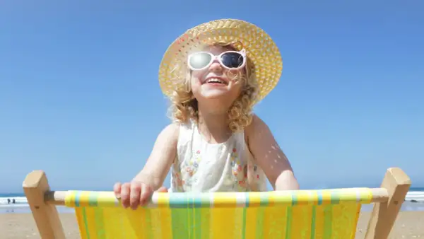 Bambina sorridente con cappello di paglia e occhiali da sole, seduta su una sdraio. Posso usare la crema solare per bambini per proteggerla dal sole.