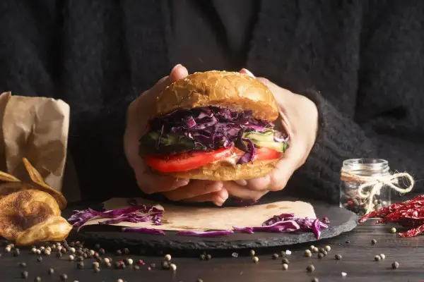 Ręce trzymają soczystego burgera z czerwoną kapustą, pomidorem i ogórkiem. Idealny przykład, jak składać burgera.