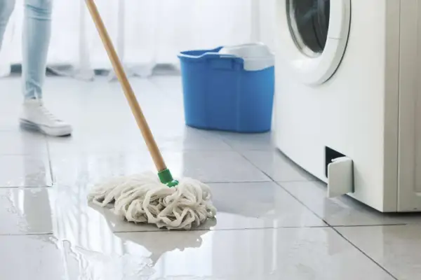 Eine Person wischt Wasser auf, das aus einer Waschmaschine austritt. Ein blauer Eimer steht bereit.