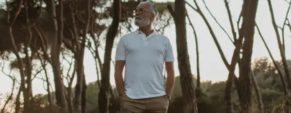Älterer Mann mit Bart in weißem Polo und brauner Hose, steht im Wald. Die polo farben sind klassisch und zeitlos.