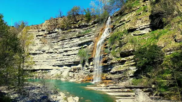 Cascate di Moraduccio: una cascata scende da una parete rocciosa stratificata, formando un arcobaleno nell'acqua turchese sottostante.