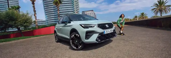 Jasnoszary SEAT Arona, jeden z najnowszych seat modele, zaparkowany obok młodej kobiety na deskorolce.