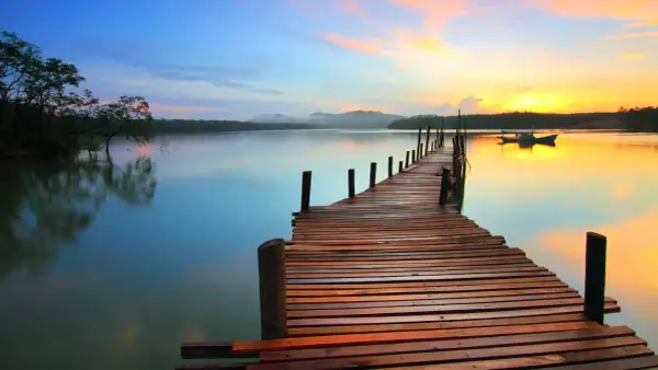 A wooden pier stretches into a calm lake at sunset, showcasing great depth of field photography. The sky transitions from blue to vibrant orange, reflecting on the water.
