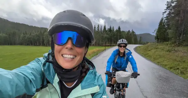 Dos ciclistas disfrutan de la ruta EuroVelo 6, con un paisaje verde y montañoso bajo un cielo nublado.