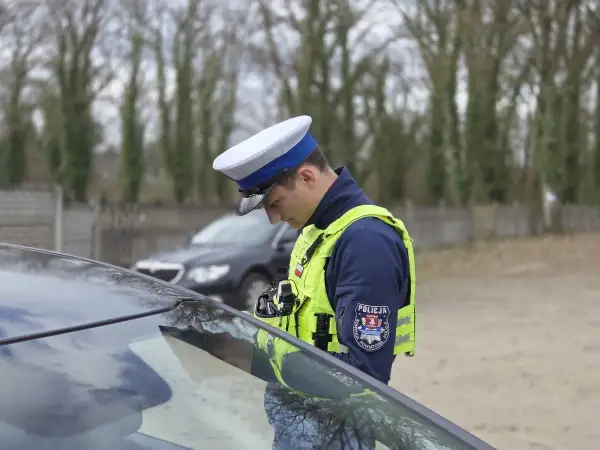 Policjant w kamizelce odblaskowej i czapce, pochylony nad maską samochodu, stosuje zasadę ograniczonego zaufania.