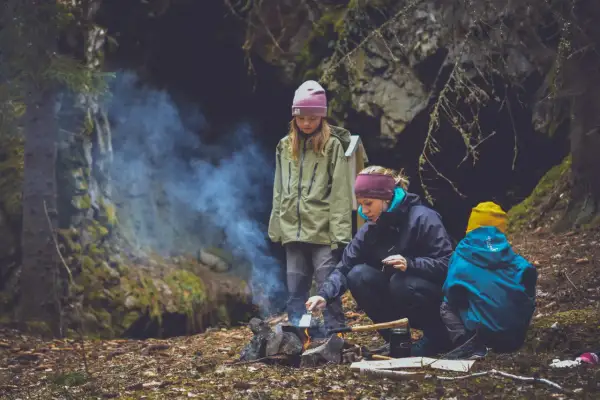 Familj lagar mat ute vid en lägereld i skogen. Rök stiger från elden där en kvinna steker något.