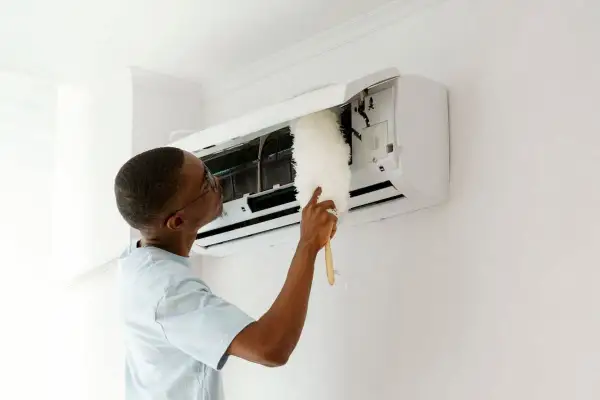 Un homme nettoie son climatiseur avec un plumeau. Un entretien régulier est essentiel pour savoir entretien climatisation tous les combien.