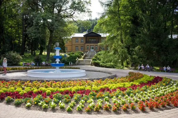 Uzdrowisko z fontanną i kolorowymi rabatami kwiatowymi przed budynkiem sanatorium.