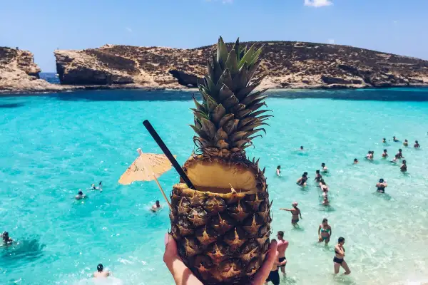Koktajl z ananasa na plaży na Malcie. Woda jest turkusowa, a na horyzoncie widać skaliste klify. Ceny na Malcie są przystępne.