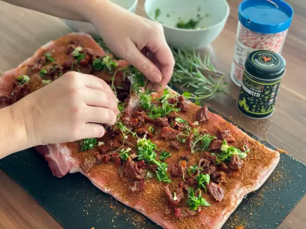 Hände belegen rohes Schweinefleisch mit Kräutern und getrockneten Tomaten für Porchetta.