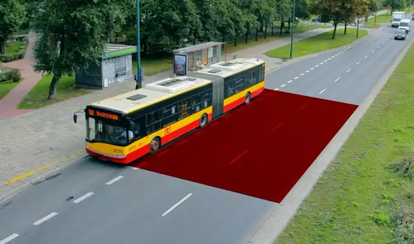 Czerwony obszar na drodze pokazuje bezpieczną odległość między samochodami w terenie zabudowanym, ilustrując przestrzeń dla autobusu.