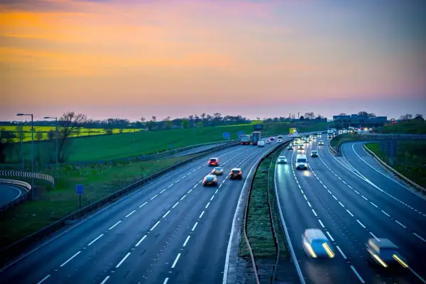 Autostrada z ruchem samochodowym o zachodzie słońca, z zielonymi polami w tle.