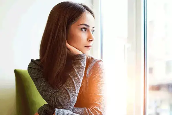 Donna pensierosa guarda fuori dalla finestra, preoccupata per un ciclo in ritardo e come farlo venire.