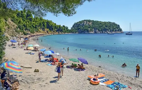 Słoneczny dzień na plaży w Chalkidiki. Ludzie opalają się, pływają w turkusowej wodzie, a kolorowe parasole rzucają cień.