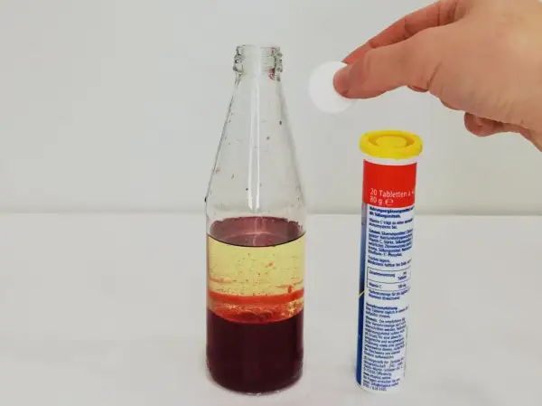 Eine Hand lässt eine Tablette in eine Flasche mit bunten Flüssigkeiten fallen, um eine Lavalampe selber zu machen.
