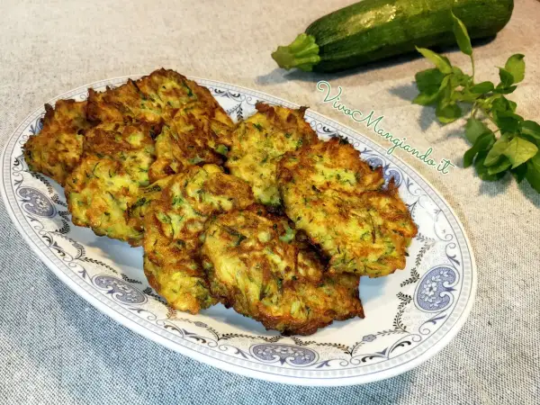 Frittelle di zucchine dorate e croccanti, servite su un piatto decorato. Accanto, una zucchina fresca e un rametto di erbe aromatiche.