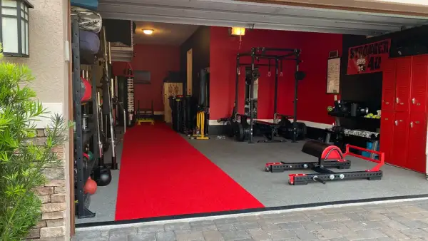 A fully equipped garage gym with a red carpet runner, weight racks, a punching bag, and a "Stronger 40" banner, ready for some serious garage boxing.
