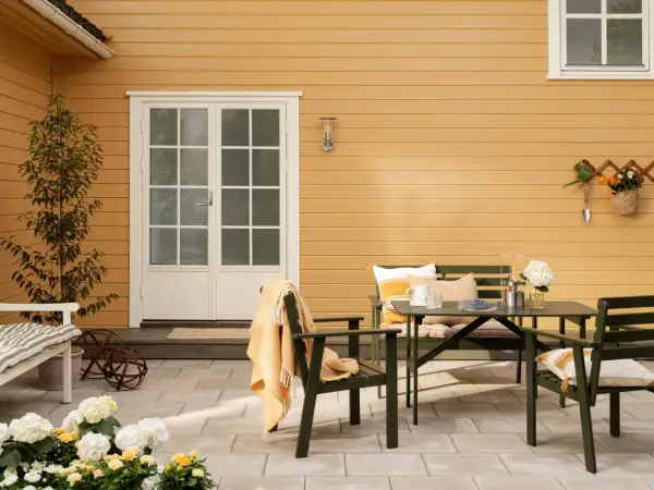 Ett gult hus med svart tak. En uteplats med gröna möbler, blommor och en orange pläd.