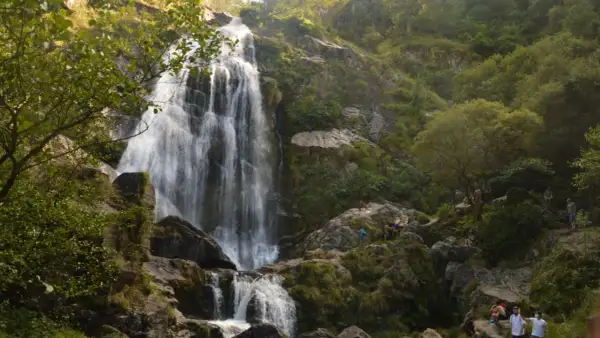 La majestuosa cascada de Belelle cae en cascada por rocas cubiertas de musgo, con gente disfrutando del paisaje y el agua.