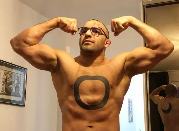 Muskularny mężczyzna o arabskich rysach, w okularach, prezentuje swoje bicepsy. Na klatce piersiowej ma tatuaż w kształcie pierścienia.