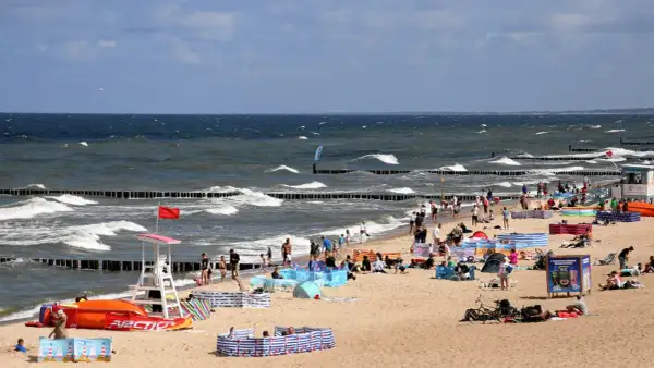 Plaża nad Bałtykiem pełna ludzi, fal i parawanów. Widać ratownika, sprzęt wodny i tablicę informacyjną. Ceny nad morzem pewnie wysokie, ale widok wart zachodu.