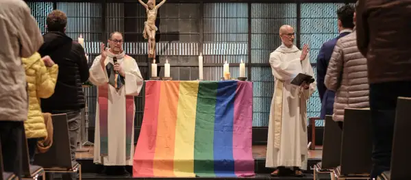 Zwei Geistliche stehen vor einem Altar mit Regenbogenflagge. Ein Kreuz hängt im Hintergrund. Die Kirche feiert die gleichgeschlechtliche Ehe.