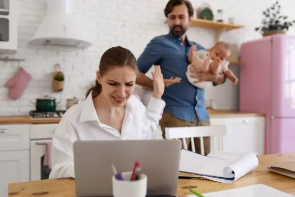 Zestresowana kobieta przy laptopie, mąż z dzieckiem w tle. Czy przerwa na karmienie piersią do jakiego wieku dziecka jest możliwa w takiej sytuacji?