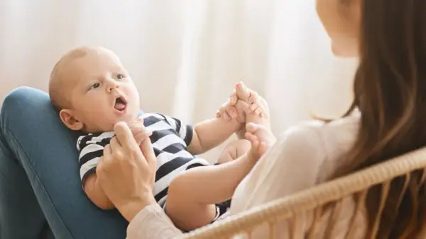 Ein Baby mit offenem Mund liegt auf dem Schoß seiner Mutter. Es hält ihre Hände. Bald sagt das Baby "Mama".