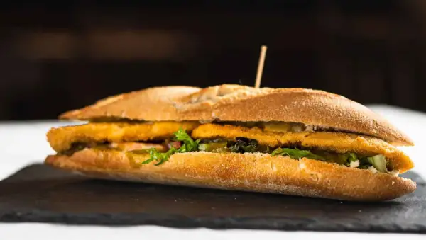 Un bocadillo caliente con rebozado crujiente, lechuga fresca y pepinillos, servido en pan rústico.