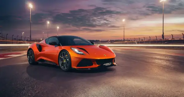 Pomarańczowy Lotus Emira 420 Sport na torze wyścigowym o zachodzie słońca, z rozmazanymi światłami w tle.