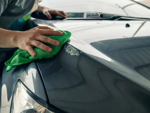 Manos puliendo el capó de un coche gris con un paño verde, mostrando cómo quitar arañazos del coche.
