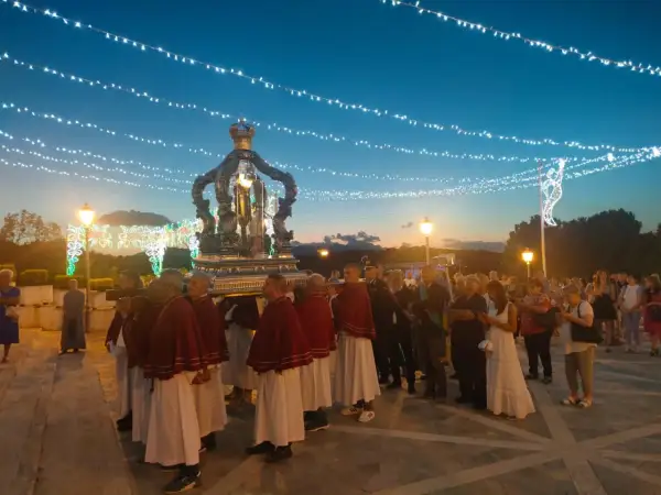 Processione serale con un carro decorato, illuminato da luci, che porta una statua. La folla osserva, mentre luci festive adornano il cielo.