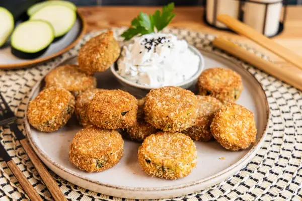 Knusprig panierte Zucchini-Taler auf einem Teller, serviert mit einem Dip und garniert mit Petersilie. Ein leckerer Snack!